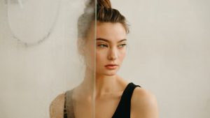A contemplative woman gazes away, standing behind a foggy glass surface indoors.