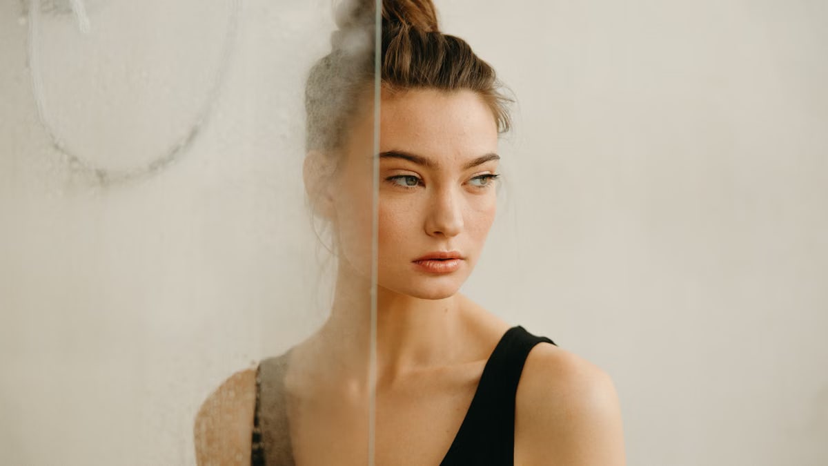A contemplative woman gazes away, standing behind a foggy glass surface indoors.